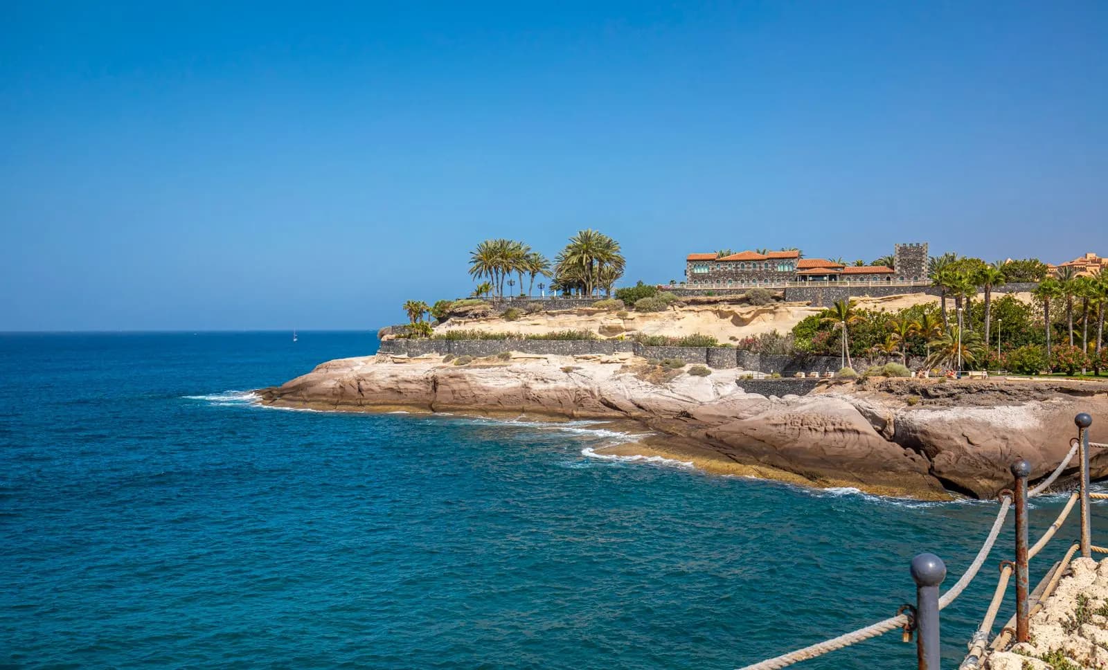Rocky headland with palm trees and a stone building above deep-blue sea on the Costa Adeje coast, Tenerife, under a clear sky