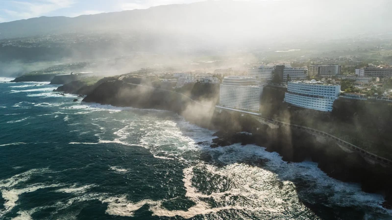 Aerial view of the Puerto de la Cruz coastline, with hotels on dark cliffs, surf and mist drifting over the green north of Tenerife