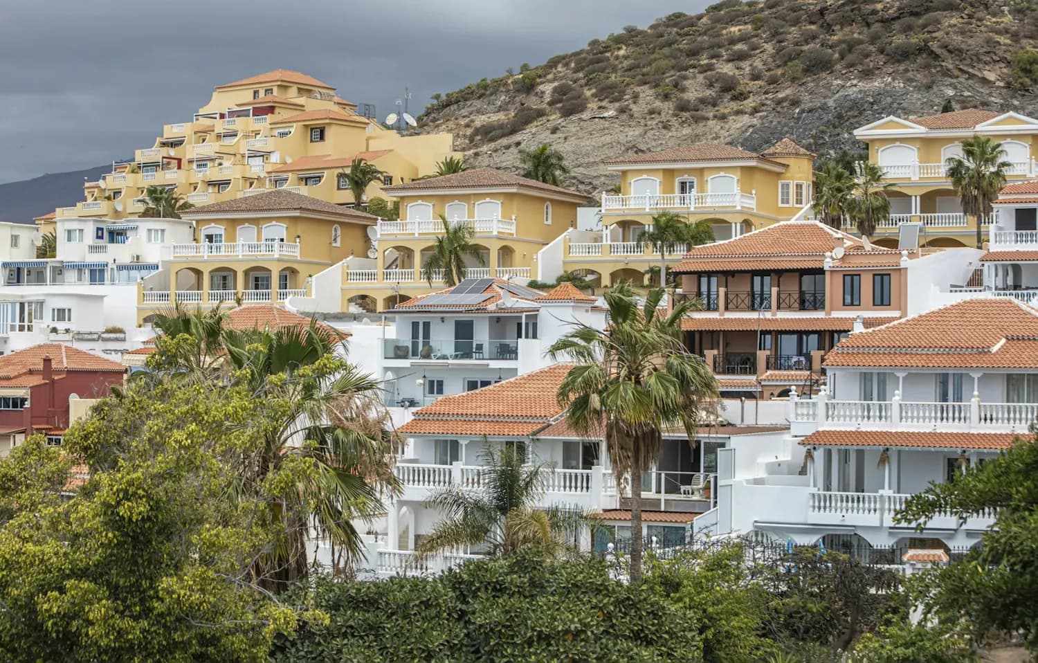 Yellow and white holiday villas with terracotta roofs, balconies and palm trees on a hillside in Costa Adeje, Tenerife, under a grey sky