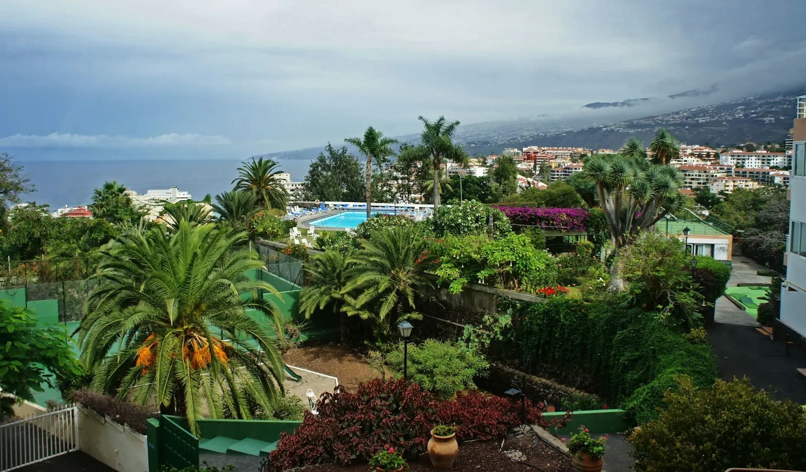 View over a holiday complex garden and pool towards the apartment blocks of Puerto de la Cruz and the sea, under grey cloud