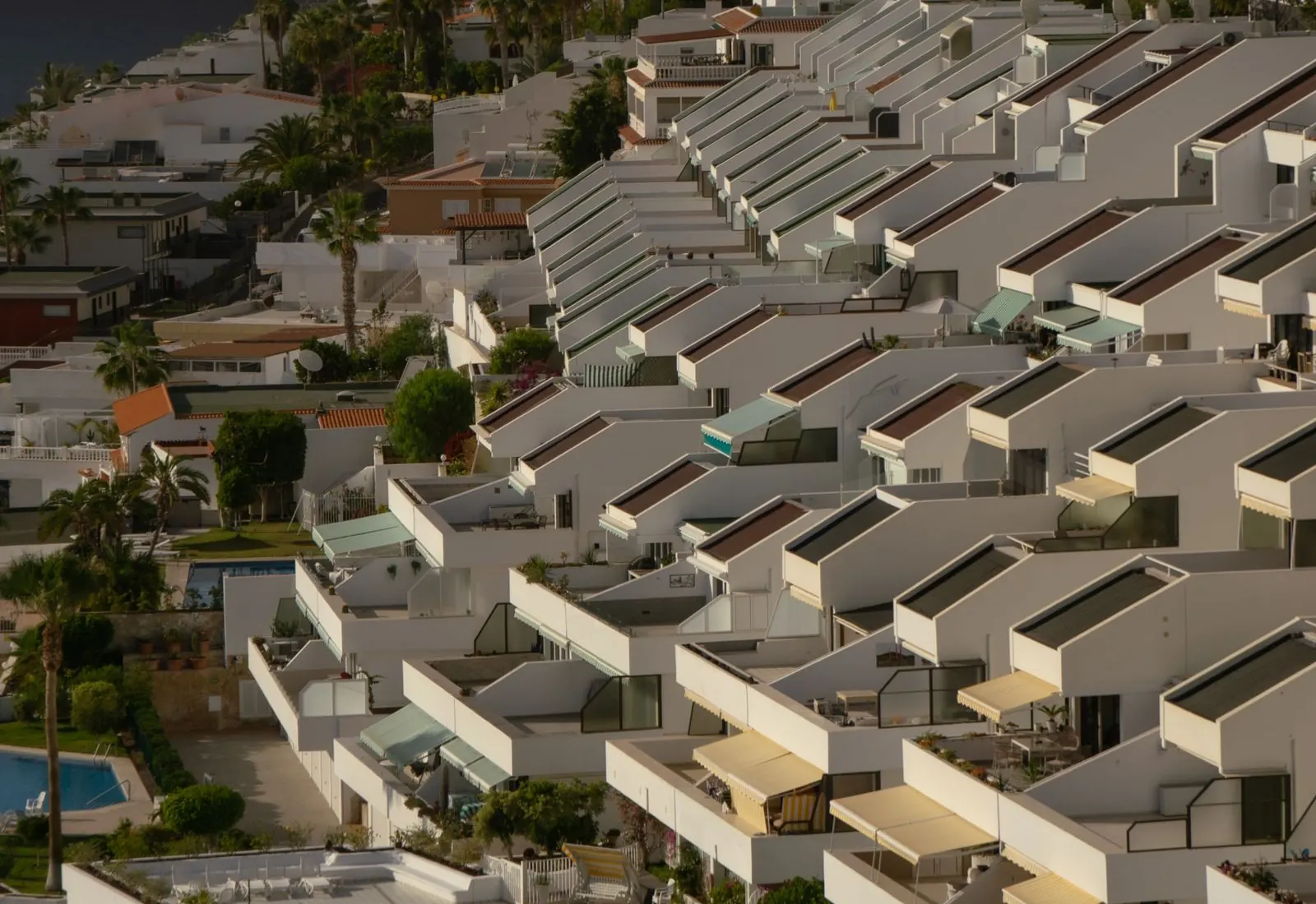 Rows of white terraced holiday apartments with awnings, gardens and a shared pool on a slope in Los Gigantes, Tenerife