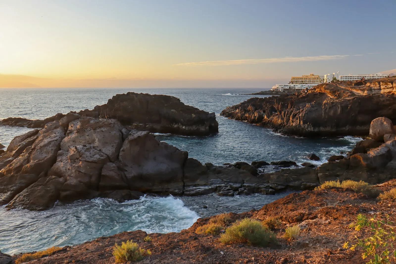 Sunset over a rocky volcanic cove on the Adeje coast, with hotels on the clifftop and the Atlantic beyond