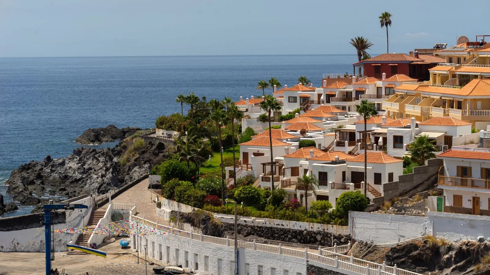Low-rise white villas with orange tiled roofs and palm trees above a rocky shore in Tenerife