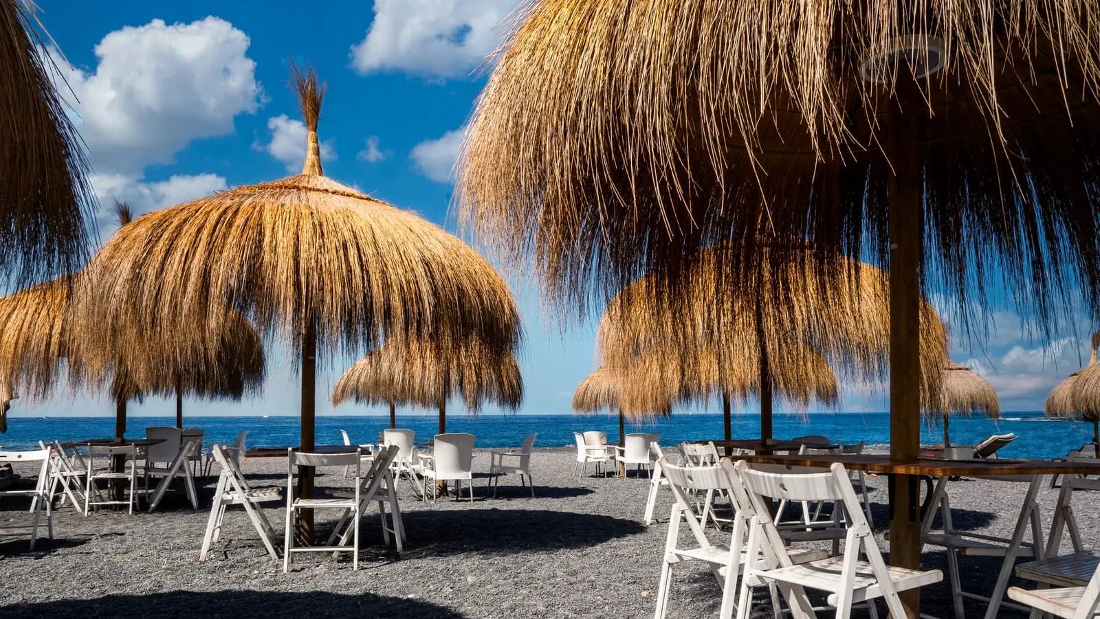 Ombrelloni di paglia e sedie bianche su una spiaggia di sabbia scura ad Adeje, sulla soleggiata costa sud di Tenerife, sotto un cielo azzurro con qualche nuvola