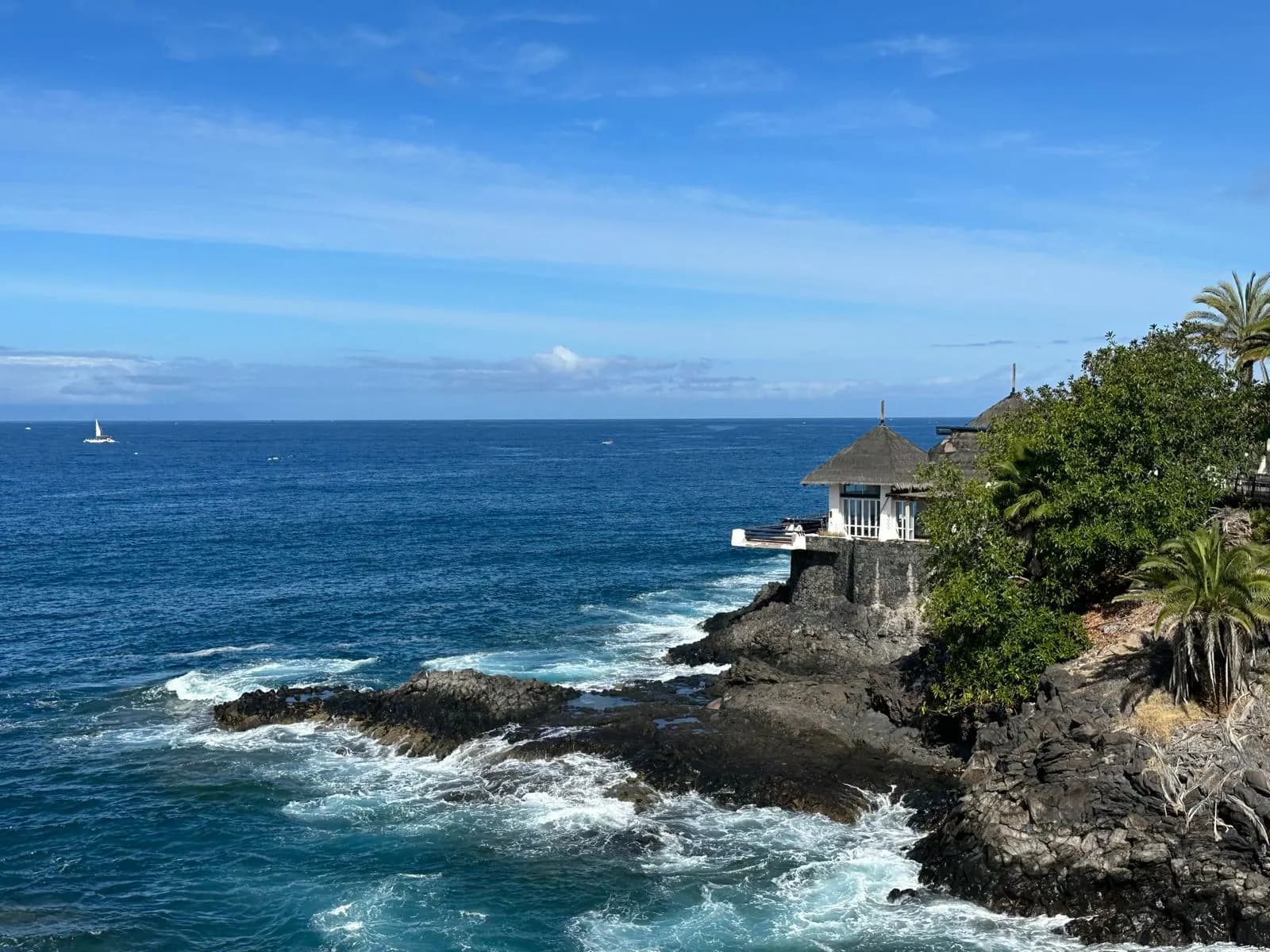 Waves breaking over dark volcanic rocks on the Costa Adeje shoreline, with a thatched seaside building and palm trees