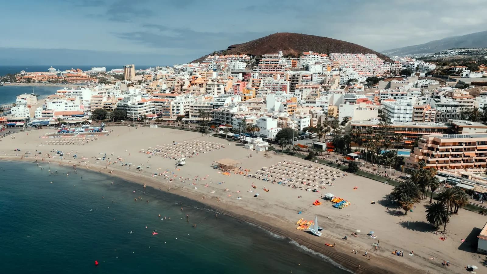 Aerial view of the sandy beach in Los Cristianos with rows of sunbeds, the white town behind and a brown volcanic hill