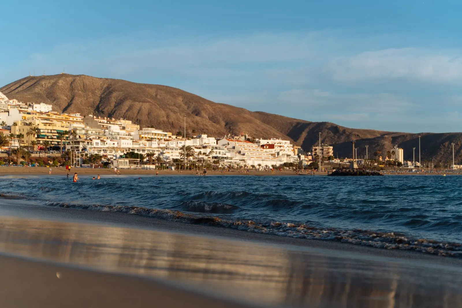 Waves on the sand of a Los Cristianos beach in late-afternoon light, with white seafront apartments and the Montaña de Guaza behind