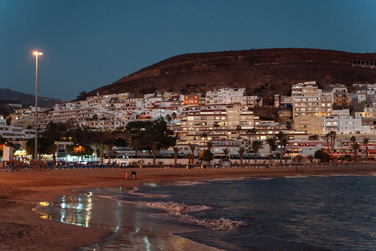 Los Cristianos beach at dusk, with lit apartment blocks climbing the hill behind the palm-lined seafront, Tenerife