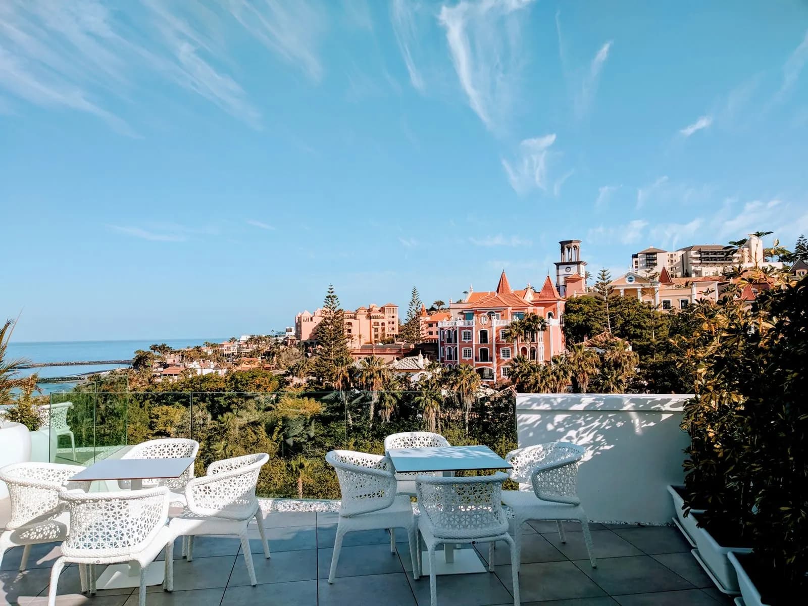 Hotel terrace with white chairs in Costa Adeje where the sea shows only as a strip on the left, beyond rooftops and palm trees