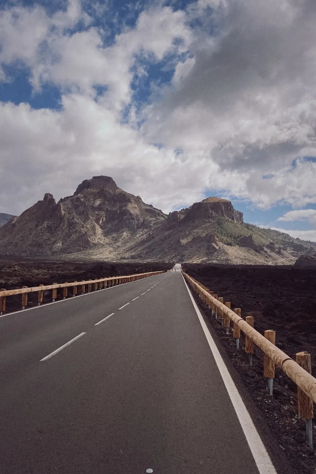Strada deserta con guardrail in legno tra colate di lava scura verso le cime rocciose del Parco Nazionale del Teide