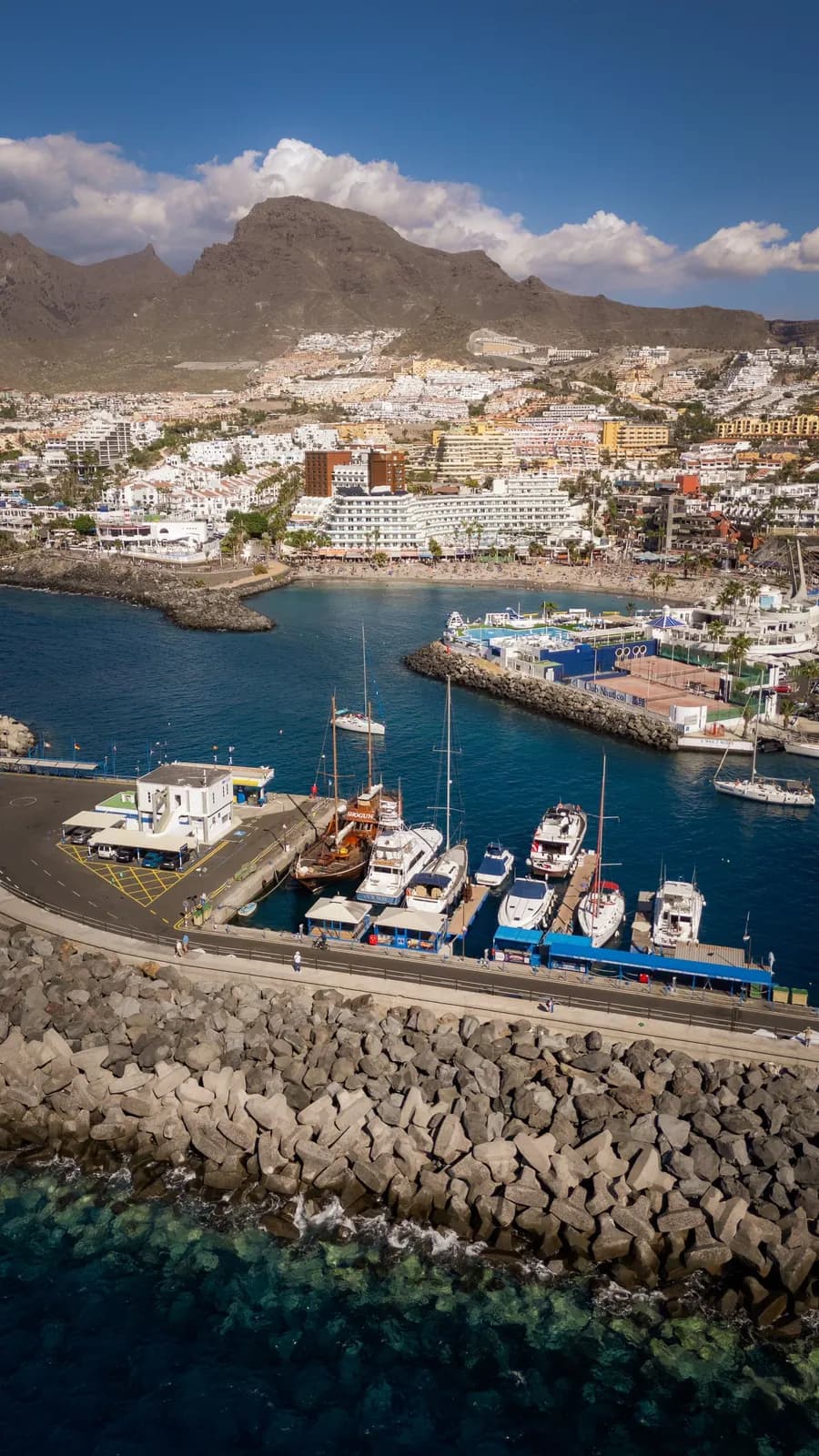 Aerial view of Los Cristianos harbour in Tenerife, with moored boats, the town beach and hillside apartments below the mountains