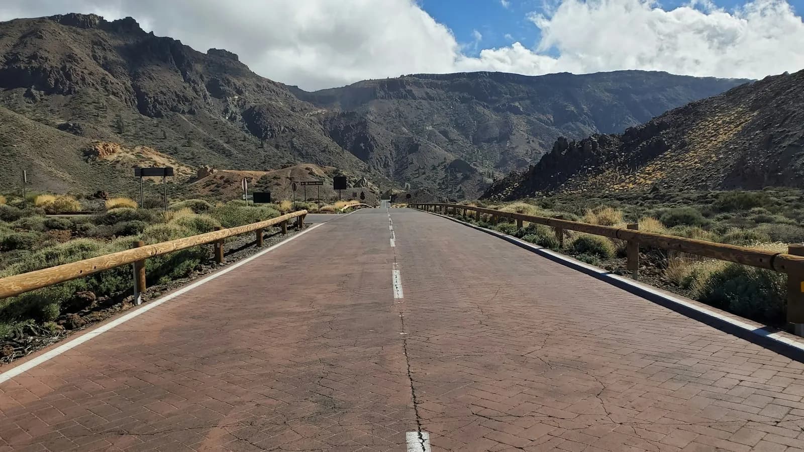 Paved road crossing the volcanic landscape of Teide National Park, with wooden guardrails and crater walls under a cloudy sky