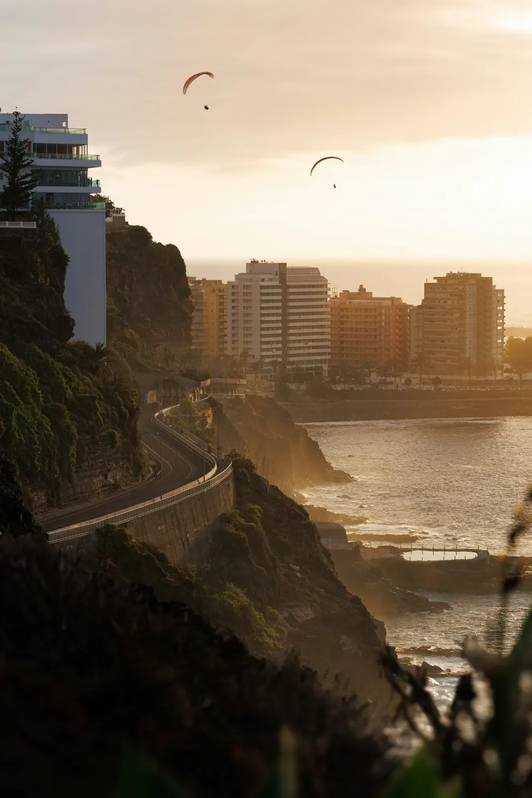 Puerto de la Cruz on Tenerife's north coast at sunset, with a cliff road, seafront apartment blocks and paragliders overhead
