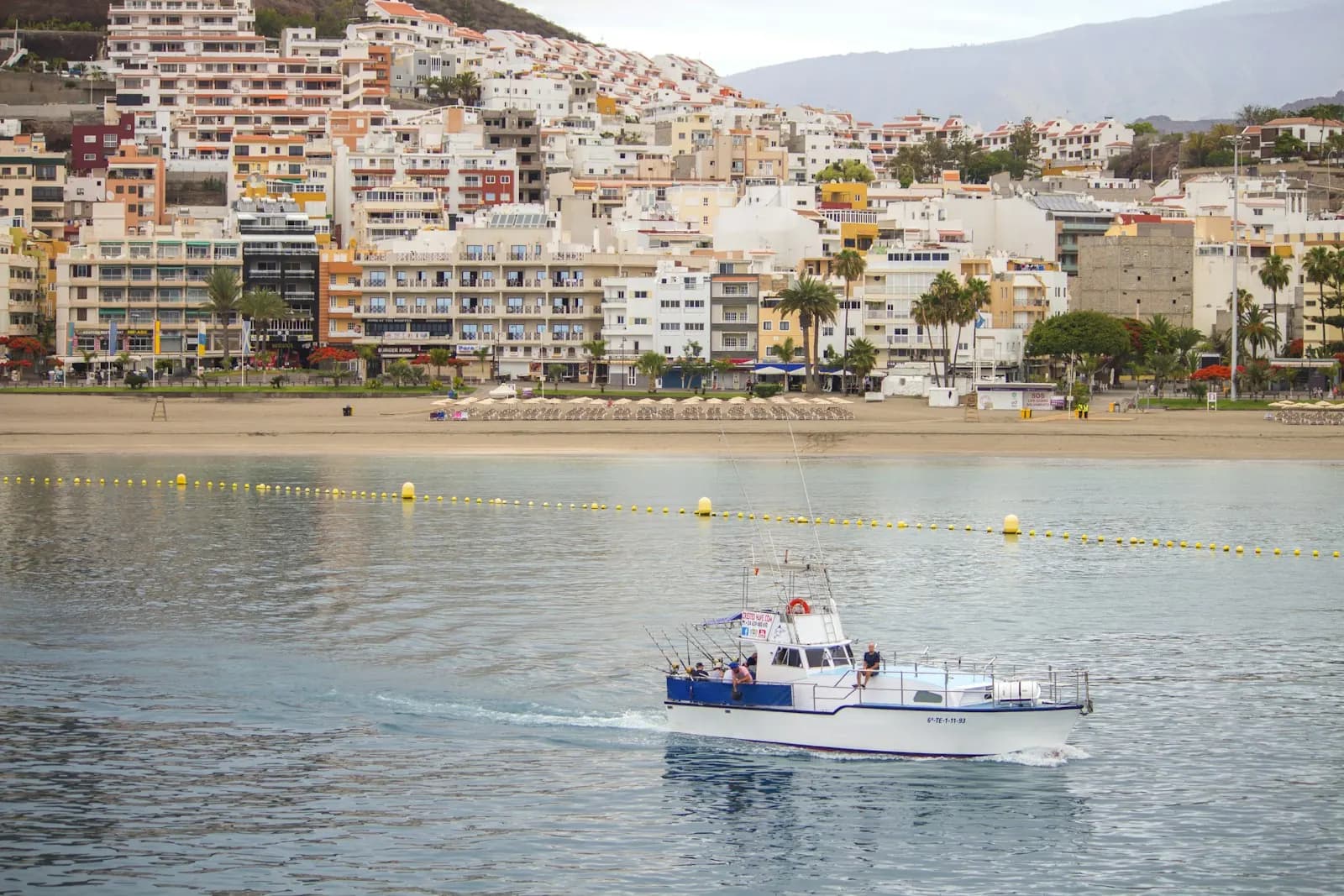 Playa de Los Cristianos with its sandy beach and hillside apartments, and a small fishing boat crossing the calm bay