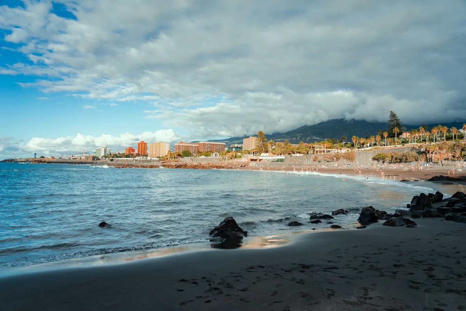 Black-sand beach on the Puerto de la Cruz seafront with volcanic rocks, palm gardens and hotels under a cloudy sky