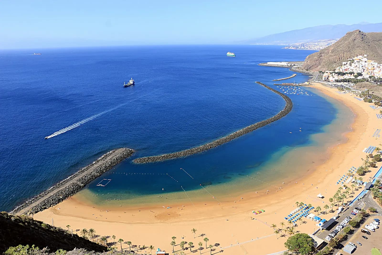 La playa de Las Teresitas vista desde lo alto, con arena dorada, el dique curvo, el agua en calma y el pueblo de San Andrés